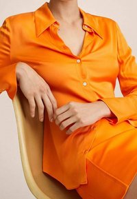 Blusa di raso arancione brillante con colletto, caratterizzata da dettagli con bottoni e maniche lunghe, indossata con una gonna abbinata, posata su una sedia.