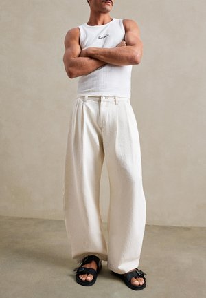 Homme aux bras croisés portant un haut blanc sans manches, un pantalon large crème et des sandales noires à boucle, debout à l'intérieur.