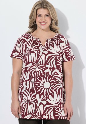 Uśmiechnięta kobieta z falującymi blond włosami, ubrana w bordową bluzkę z krótkim rękawem ozdobioną białymi abstrakcyjnymi wzorami dłoni i roślin.