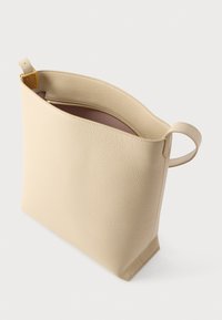 Beige handtas van getextureerd leer met een bovenrits, rechthoekige bodem en een lusvormig handvat, op een witte achtergrond.