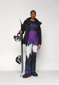 Snowboarder i svart jakke, lilla og blå mønstrede bukser, holder et snowboard. Funksjoner inkluderer justerbare stropper og teksturert stoff.