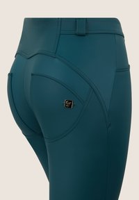 Leggings teal a vita alta con cuciture curve e un piccolo logo dorato sul retro. Materiale liscio ed elastico senza motivi visibili.