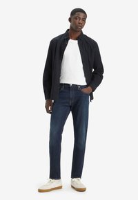 Camisa negra de botones sobre una camiseta blanca, combinada con jeans oscuros y zapatillas de tono claro. Diseño simple y ajustado con acentos mínimos.