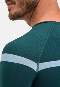 Zielona sportowa koszulka z teksturowanym wzorem, z jasnoszarym poziomym paskiem na wysokości klatki piersiowej i gładkimi szwami na krawędziach.