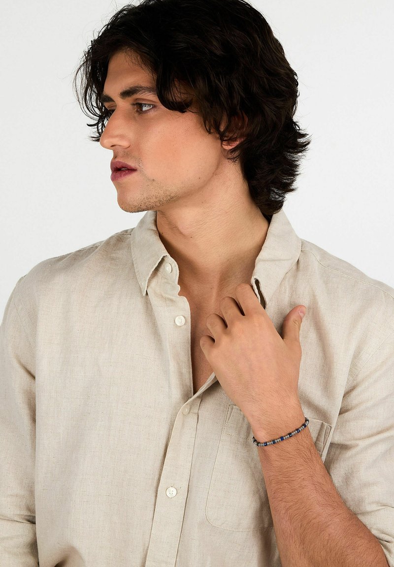 Camicia di lino di un leggero beige con colletto Button-down, dotata di taschino. Indossata con un semplice braccialetto di perline al polso.