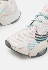 Scarpe atletiche leggere con una parte superiore bianca testurizzata, dettagli turchesi e una suola multicolore maculata. Presentano elementi di design a motivi.