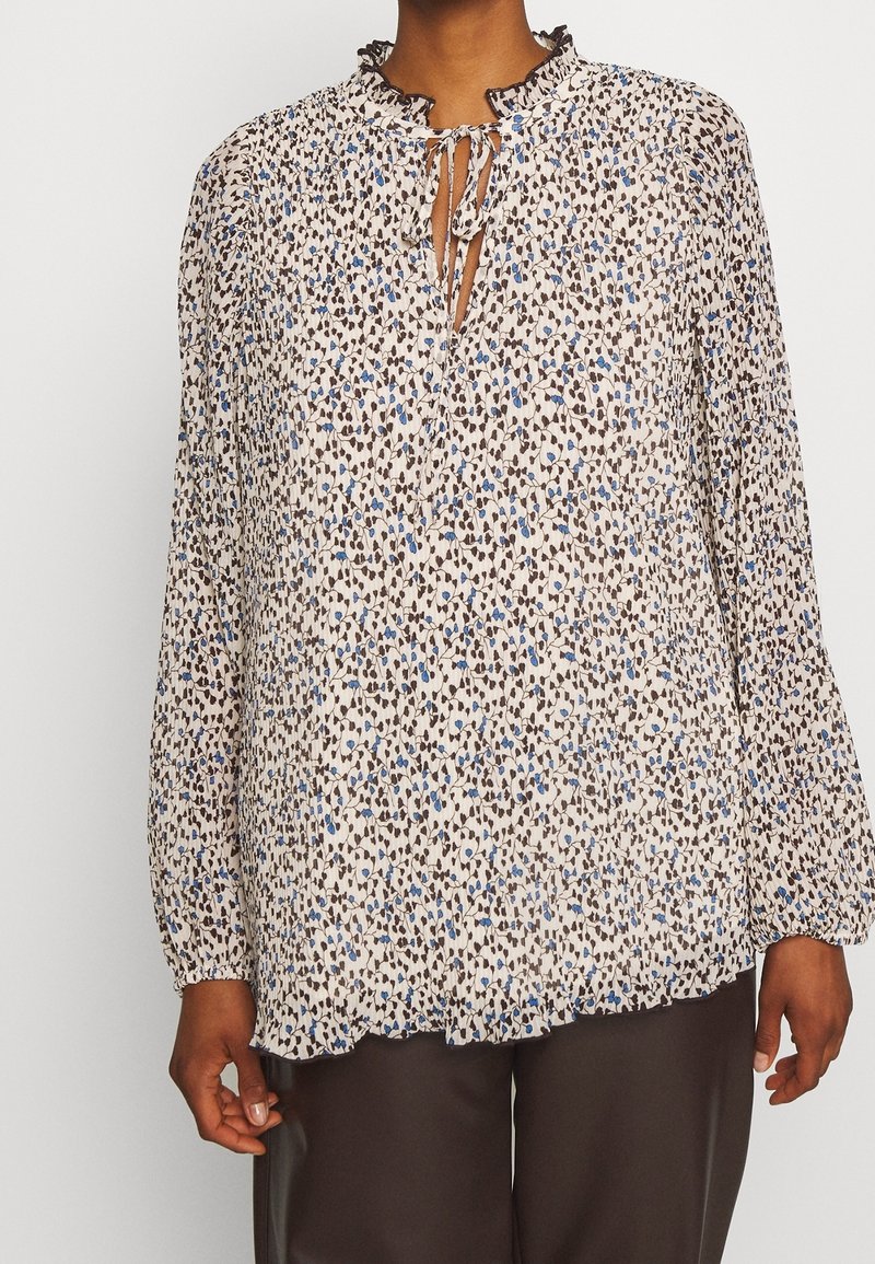 Femme portant un chemisier à manches longues avec un petit motif floral bleu et noir, col à nouer et pantalon marron.