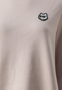 T-shirt beige chiaro realizzato in morbido cotone, con una patch a forma di gatto in bianco e nero con occhiali da sole sulla parte anteriore. Texture liscia.