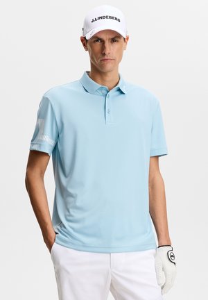Lyseblå polo med krave, korte ærmer og diskrete stribede detaljer på ærmerne. Båret sammen med hvide bukser og en hvid kasket.