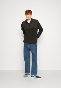 Svart fleece-pullover med kvart-glidelås krage og et fargeblokkert design, paret med avslappede blå denimjeans og svarte joggesko.