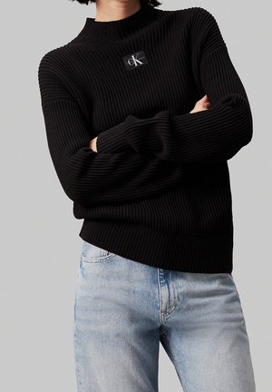 Persoon draagt een geribde zwarte Calvin Klein trui met logopatch op de borst, lichtblauwe jeans, armen gekruist, grijze achtergrond.