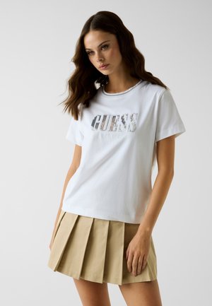 Jeune femme aux cheveux bruns ondulés porte un t-shirt blanc avec le logo Guess et une mini-jupe plissée beige, debout devant un fond clair uni.