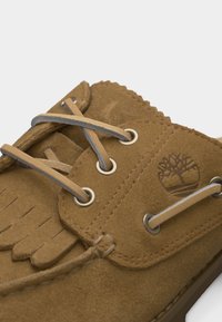 Mocassin en daim marron avec des lacets en cuir passés dans des œillets métalliques et un logo d'arbre estampé sur le panneau latéral.