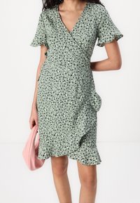 Grön omlottklänning med mönster av svarta och vita blad, korta volangärmar, asymmetrisk nederkant och en mjuk textur. Kombinerad med en rosa handväska.