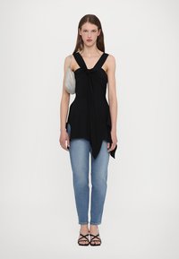 Μαύρο αμάνικο τοπ με λεπτομέρεια κόμπου μπροστά, συνδυασμένο με ανοιχτό μπλε τζιν και μαύρες σαγιονάρες με λουριά; κρατώντας μια ασημένια τσάντα.