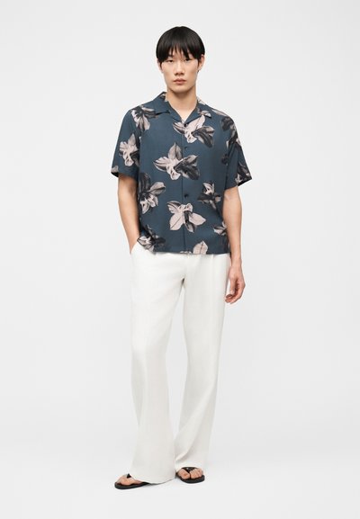 Jeune homme portant une chemise à manches courtes florale bleu foncé, un pantalon blanc à jambes larges et des sandales noires, debout devant un fond uni.