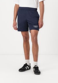Marineblaue Sportshorts aus atmungsaktivem Material, mit einem elastischen Bund und einer Logostickerei am linken Bein. Kombiniert mit schwarzen Sneakers.