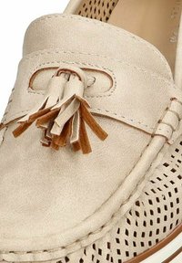 Mocasín de cuero beige con diseño perforado, terminado con un borlas decorativa y acentos en tono marrón claro. Textura suave, punta redondeada.