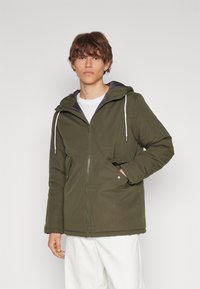 Doudoune verte olive avec une fermeture éclair à l'avant, capuche ajustable, deux poches latérales et cordons blancs. Tissu texturé avec une finition lisse.
