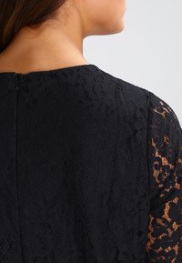 Oben am Rücken und an der Schulter einer Frau, die ein schwarzes Blumen-Spitzenkleid mit einem verdeckten Reißverschluss und halbdurchsichtigen Spitzenärmeln trägt.