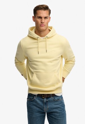 Gelber Hoodie mit großer Kängurutasche vorne, Kapuze mit Kordelzug und gerippten Bündchen. Trägt man zu blauen Jeans, präsentiert vor einem schlichten Hintergrund.