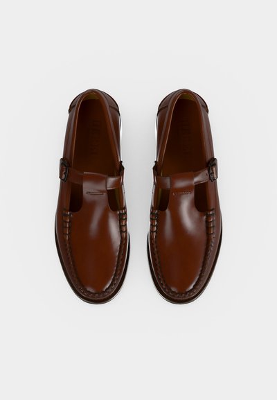 Paire de mocassins T-strap en cuir marron poli avec fermetures à boucle et coutures détaillées sur fond blanc.