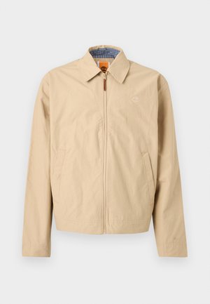 Bež jakna z zadrgo in ovratnikom, stranskimi žepi ter vezenim logotipom na prsih, z notranjo podlogo v modro karo vzorcu.