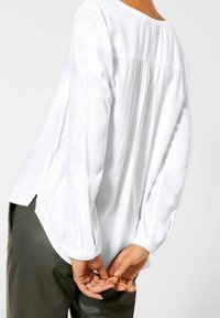 Blusa bianca a maniche lunghe con scollo rotondo, dettagli arricciati sul retro e spacco laterale, abbinata a pantaloni in pelle verde oliva.