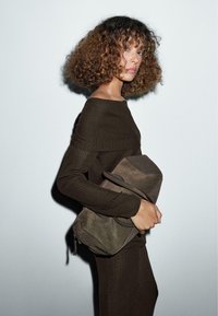 Eine Person mit lockigem Haar trägt ein dunkelbraunes geripptes Kleid und hält eine große taupefarbene Wildlederhandtasche vor einem schlichten hellen Hintergrund.