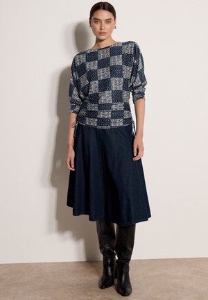 Mujer de cabello oscuro lleva una blusa de manga larga a cuadros azul marino y blanca, falda midi azul marino y botas negras hasta la rodilla, de pie contra una pared lisa.