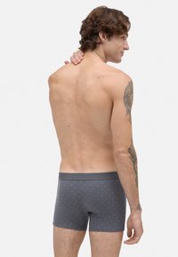 Hombre sin camiseta con cabello rizado, vistiendo calzoncillos grises con un patrón de pequeños puntos, mostrando la espalda y tatuajes en el brazo derecho y el cuello.