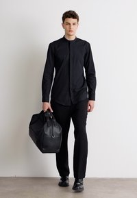Schwarzes Langarmhemd mit hohem Kragen, kombiniert mit schwarzen Hosen und Schuhen. Hält eine schwarze Leder-Duffle-Tasche, glatte Textur, minimalistisches Design.
