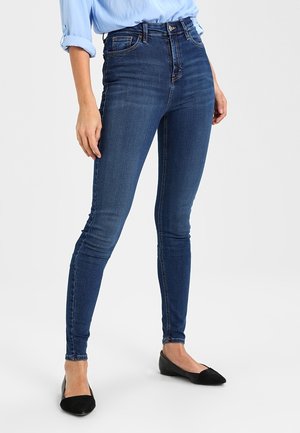 Femme portant un jean skinny bleu foncé, une chemise à manches longues bleu clair avec manches retroussées, et des ballerines noires pointues, debout sur un fond blanc.