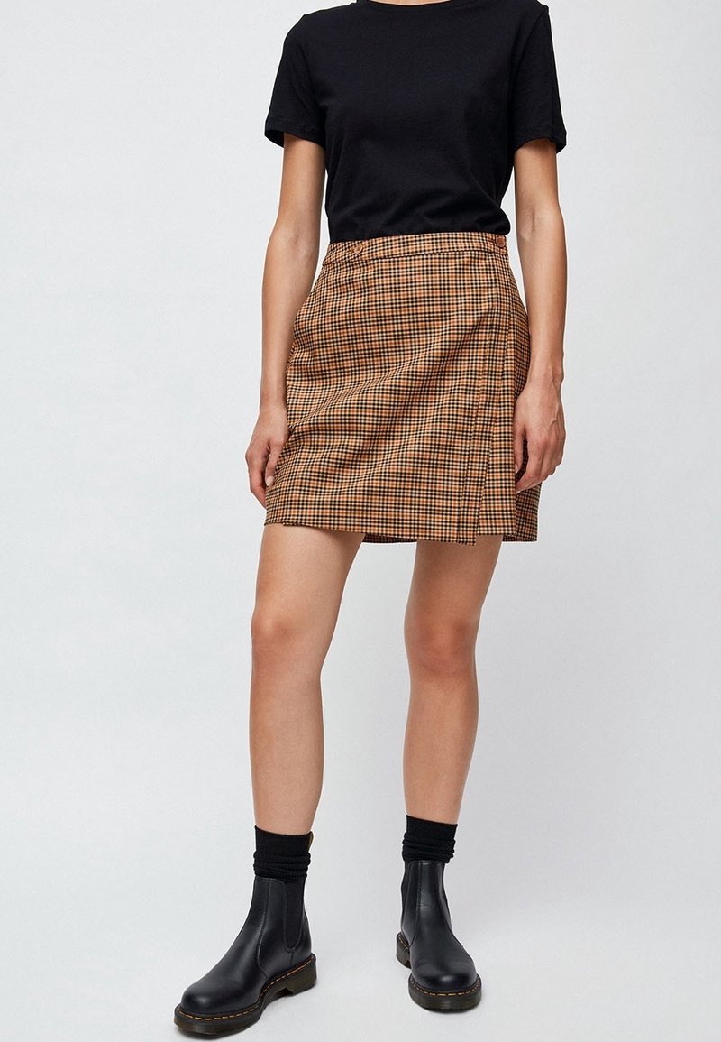 Mini-jupe écossaise marron avec une fente latérale, associée à un t-shirt noir et des bottines noires. Tissu lisse avec une légère texture.