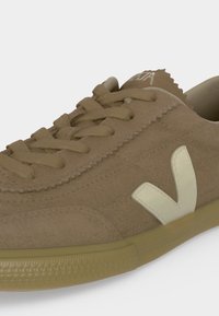 Baskets en daim beige avec lacets assortis, un logo blanc en forme de "V" sur le côté et une semelle en caoutchouc brun clair texturée.