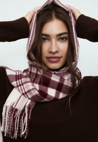 Écharpe à carreaux en bordeaux et crème, fabriquée en tissu doux. Présente des bords frangés et un motif à carreaux, portée avec un pull marron.