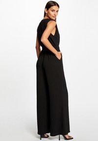 Zwarte jumpsuit met mouwloze ontwerp, elastische taille en zijzakken. Vloeiende stof en wijde pijpen passen perfect bij de minimalistische esthetiek.