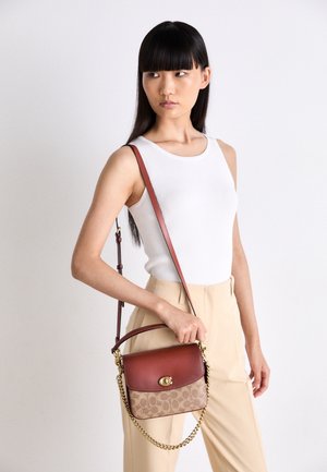 Kobieta z długimi czarnymi włosami, ubrana w biały top bez rękawów i beżowe spodnie, trzymająca brązową skórzaną torebkę z wzorem i złotym łańcuszkiem.