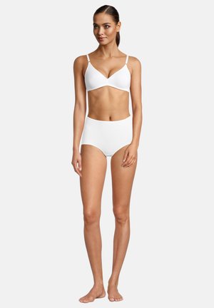 Woman with brown hair in a white bra and high-waisted briefs standing barefoot against a plain light background.