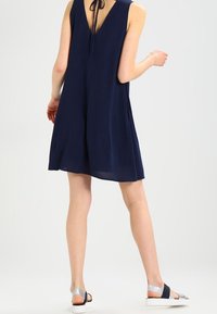 Laivastonsininen hihaton mekko, jossa on V-pääntie ja solmukiinnitys selässä, yhdistettynä hopeakiiltoisiin sandaaleihin. Mekossa on rento istuvuus ja sileä rakenne.