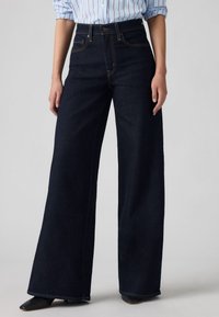 Brede donkerdenim jeans met voorzakken, hoge taille en contrasterende stiksel. Gecombineerd met een lichtblauw gestreept shirt.