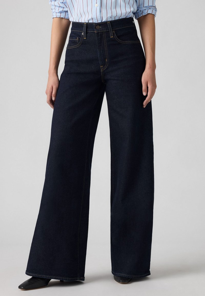 Brede donkerdenim jeans met voorzakken, hoge taille en contrasterende stiksel. Gecombineerd met een lichtblauw gestreept shirt.