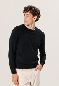 Schwarzer Pullover, Rundhalsausschnitt, gestrickte Textur, lange Ärmel, gerippte Bündchen und Saum. Wird mit hellfarbigen Hosen getragen, das Modell posiert seitlich.