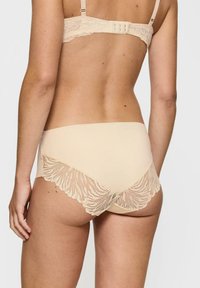 Beżowa bielizna z gładką tkaniną z przodu i koronkowym tyłem w falbanki, z wzorem przypominającym liście. Biustonosz ma regulowane ramiączka.