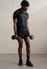 T-shirt de sport noir à manches courtes et short noir. L'individu tient deux haltères hexagonaux, portant des chaussettes et des chaussures de sport noires.