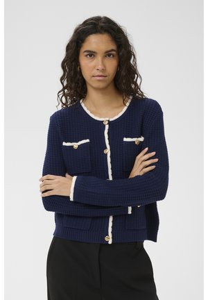 Kobieta z kręconymi włosami, ubrana w granatowy dzianinowy kardigan z białą lamówką i złotymi guzikami, stojąca ze skrzyżowanymi ramionami na tle jednolitego jasnego tła.