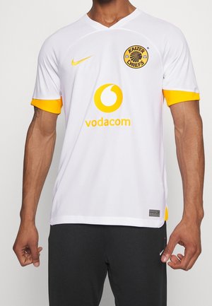 Homme portant un maillot de football blanc des Kaizer Chiefs avec un logo jaune et le sponsor Vodacom, associé à un pantalon noir Nike.
