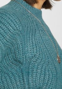 Maglione lavorato a maglia color teal con un motivo a onde testurizzato, scollatura a costine e una collana con pendente in argento. Primo piano sulla parte superiore dell'indumento.