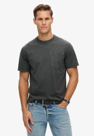 Grijze katoenen T-shirt met korte mouwen, ronde halslijn en een borstzak aan de voorkant. Draagt bij lichte blauwe gescheurde jeans en een zwarte riem.