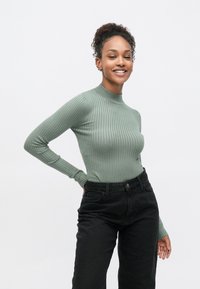 Ανοιχτό πράσινο ριπ τοπ με μακριά μανίκια και ψηλό λαιμό, σε συνδυασμό με μαύρο υψηλόμεσο τζιν με πλαϊνές τσέπες και λεπτή υφή.